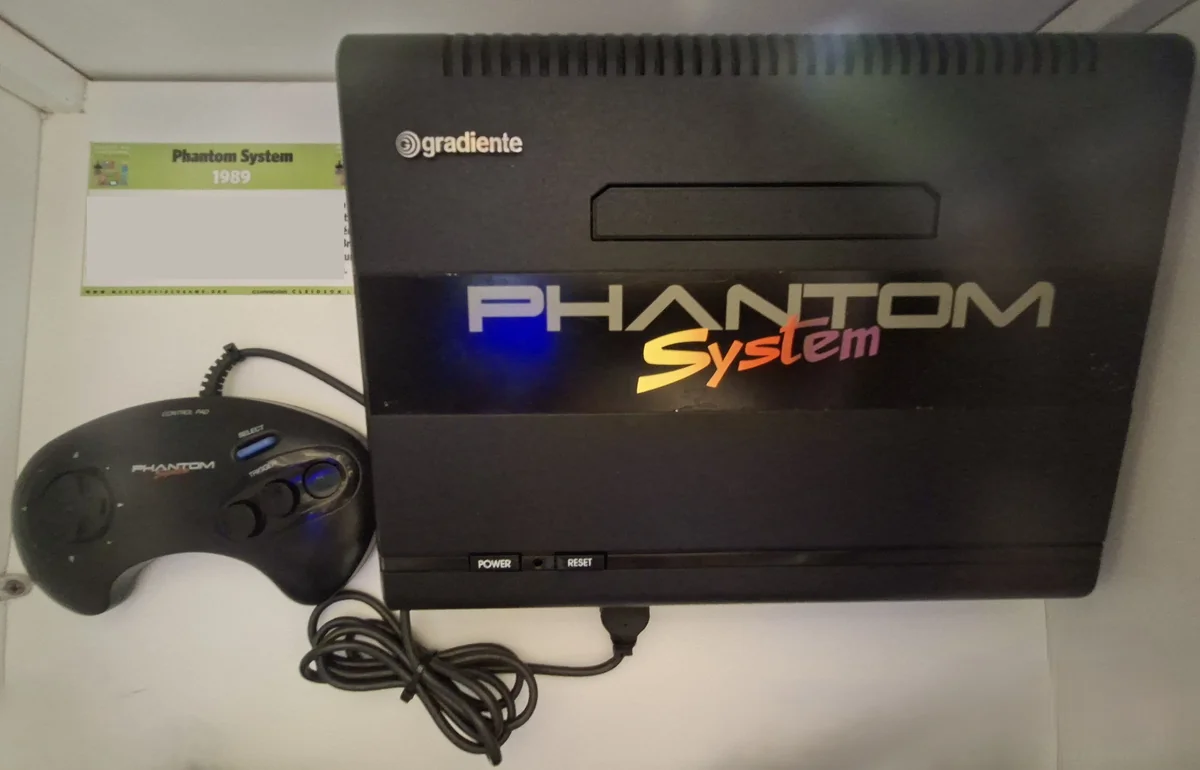 Console preto Phantom System da Gradiente ao lado do controle ergonômico, com cartaz de museu indicando Phantom System 1989